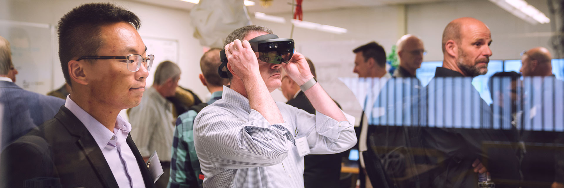 DBL members walk around a room looking at displays of Georgia Tech work. One man, in the center of the photo, wears a VR headset.