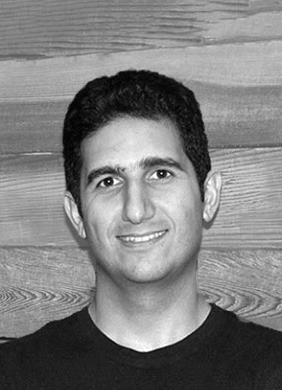 This is a grayscale portrait of Dr. Nourbakhsh, he is smiling towards the camera, wearing a black t-shirt. The background is a dark colored wood beadboard. 