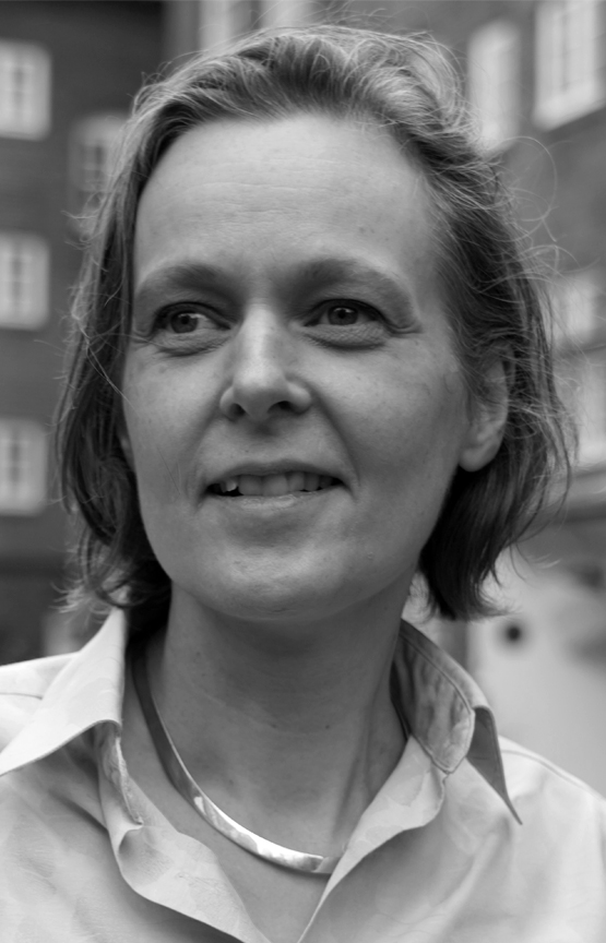 This is a grayscale portrait of Dr. Whyte. She is wearing a light coloured shirt and smiling off camera. A brick building with white window encasements is visible in the background.