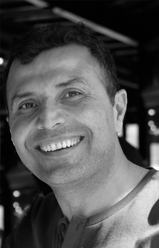 This is a grayscale portrait of Dr. Dogan. He is smiling towards the camera, wearing a dark button-up collarless shirt. There are sun patches and an intricate background with bokeh effect.