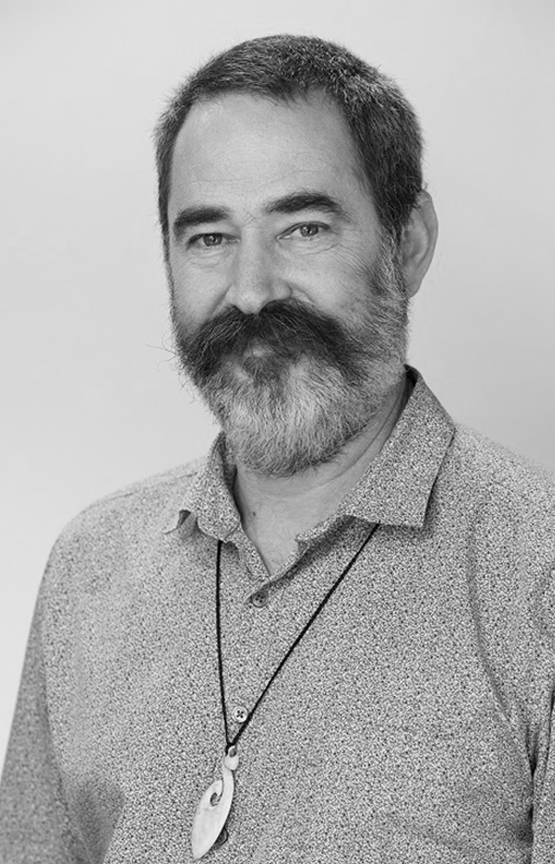This is a grayscale portrait of DR. Amor. He is smiling, wearing a perlin noise patterned shirt undone at the collar. He is also wearing a hei matau pendant.